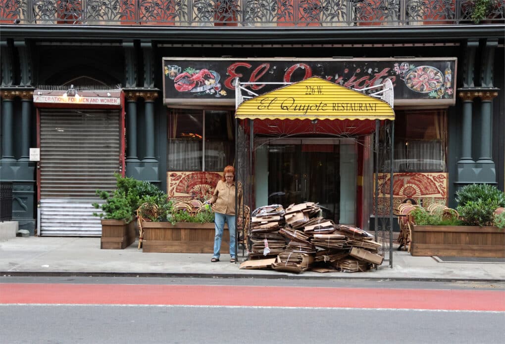 A woman stands on a sidewalk next to a restaurant awning. Several dozen cardboard boxes are bundled for recycling on the curb.