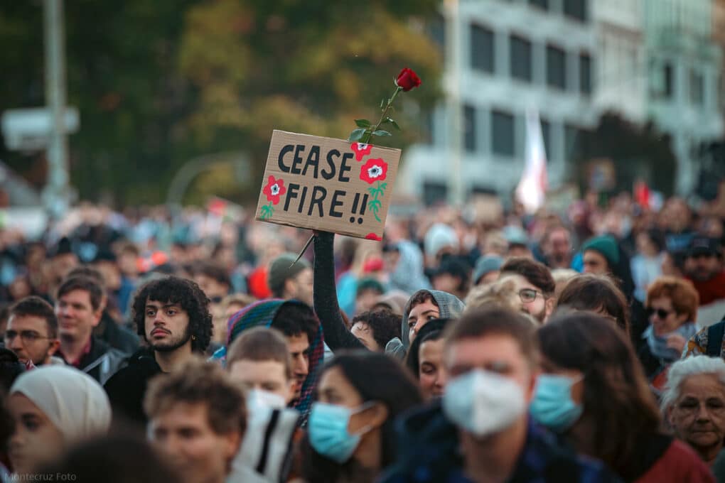A photo of many faces in a crowd. Above their heads, one person in a hooded sweatshirt holds a small piece of cardboard with "Cease Fire" painted in thick black letters and two bright red flowers. Behind the cardboard in the same hand, the person holds a red rose.