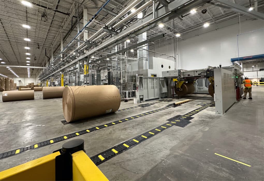 A view of a factory with various machines and huge rolls of brown paper, approximately five feet in diameter and at least twice that length.
