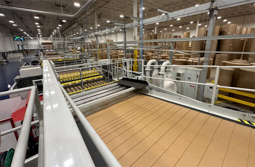 A more open view of the factory floor, with cardboard sheets coming off the end of a complicated machine. Dozens or hundreds of large brown paper rolls are stacked in the background.