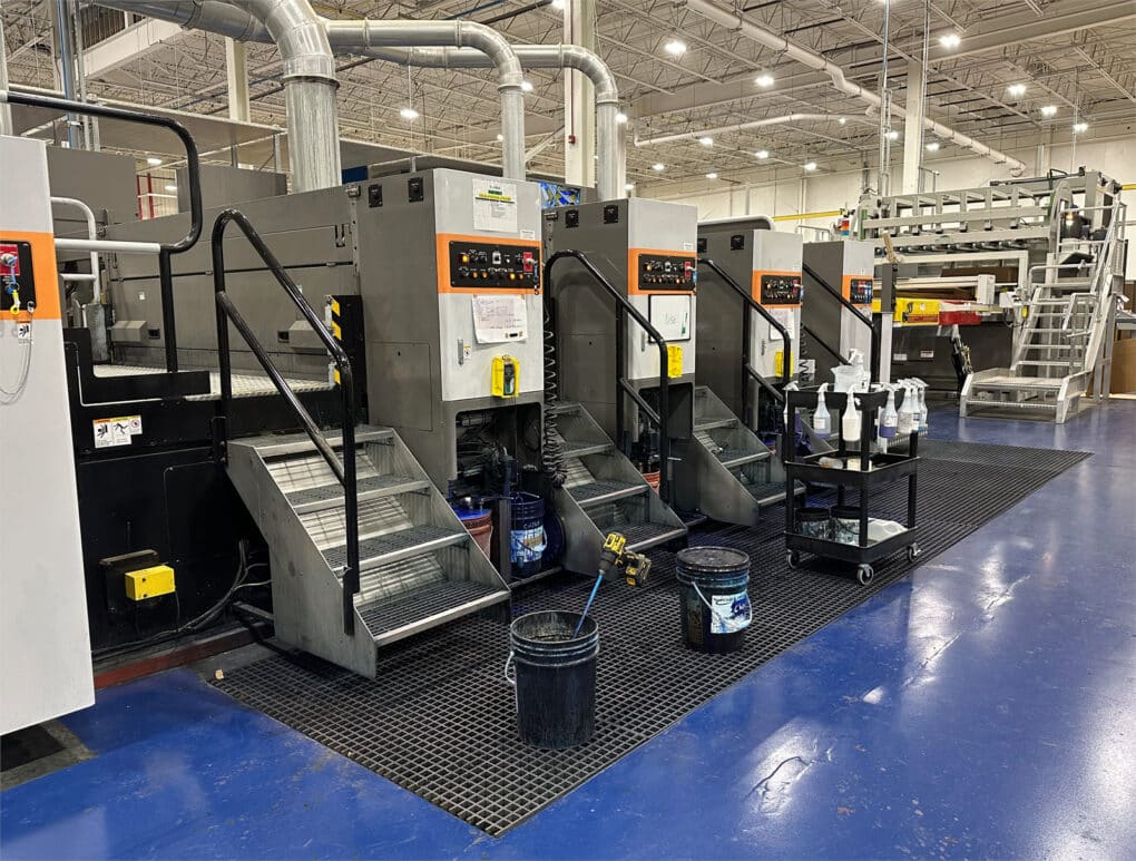 A printing press in a factory floor with four bays. There are buckets beneath each bay, and there is a cleaning station with several spray bottles.