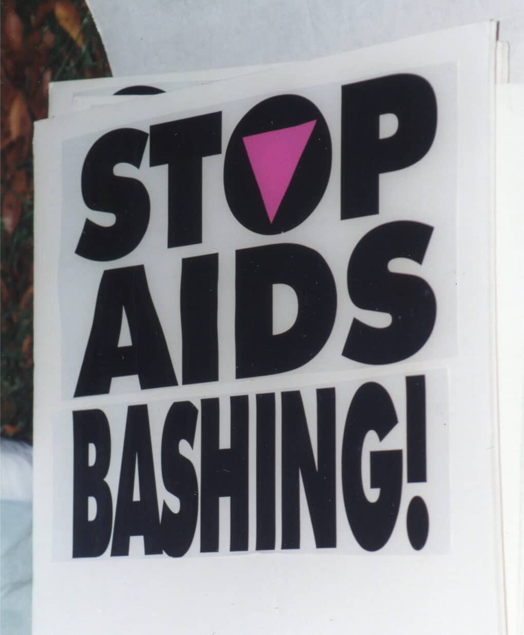 Color photograph of poster with large black san-serifed all-caps reading: STOP AIDS BASHING! A pink downward-pointing triangle forms the opening in the "O" in "STOP."