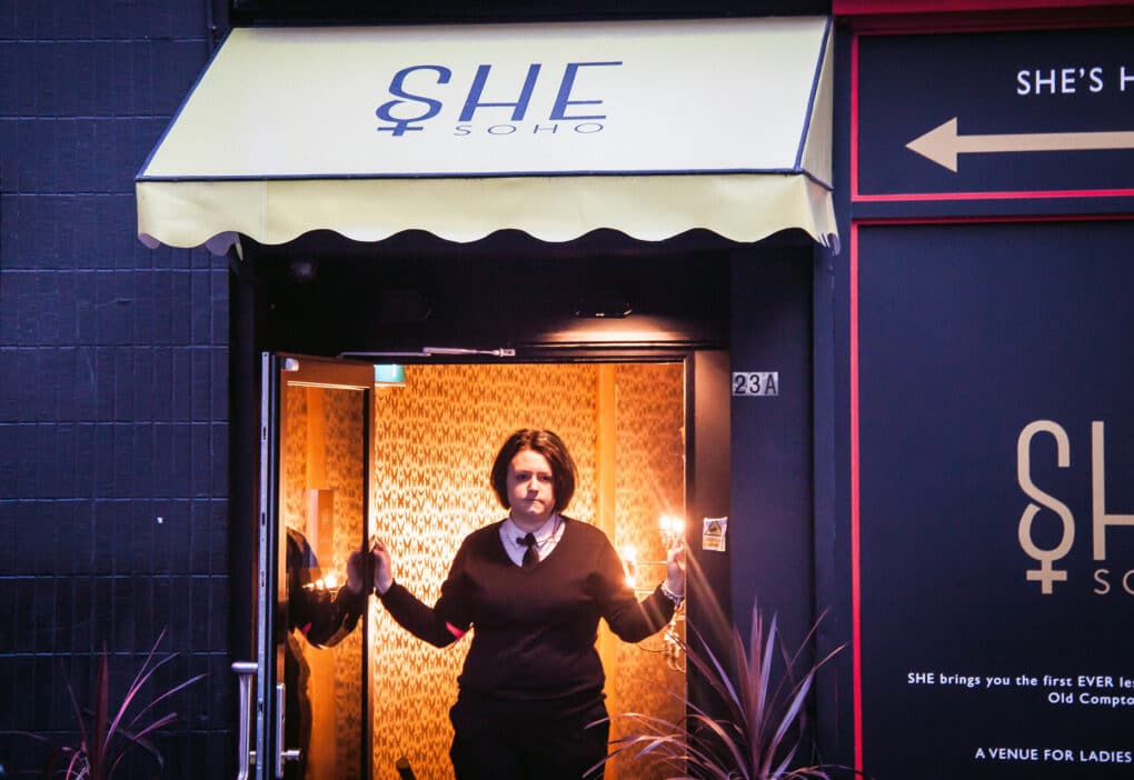 Color photograph of young white woman in collared shirt and necktie under black v-neck sweater, standing in an open doorway under an awning reading SHE SOHO.