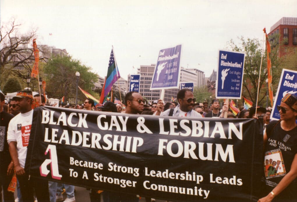 Color photograph of three black men and a black woman, carrying a black banner with white writing reading "Black Gay & Lesbian Leadership Forum Because Strong Leadership Leads To A Stronger Community." They stand in the midst of a large crowd carrying signs and banners.
