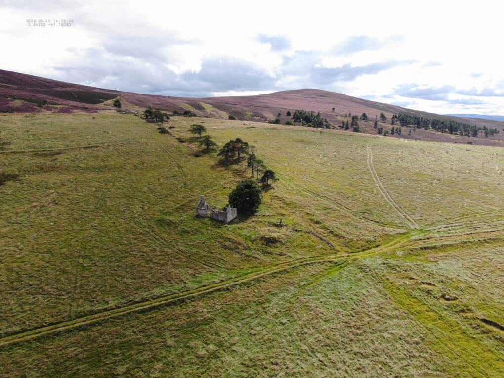 Drone photograph of roofless stone cottage at the intersection of several straight lines in a grassy field.