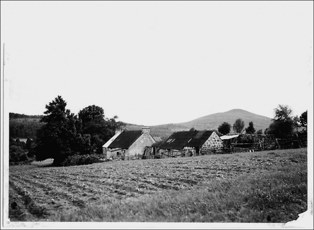 Black-and-white photograph of tidy stone cottage with stone outbuildings, cultivated field, fences, and trees.