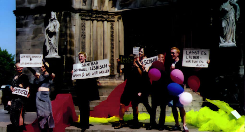 Color photograph showing group of activists, some in dresses, other in suits in a public demonstration in front of a classical church. Some hold placards; balloons and colored cloths create a festive atmosphere.