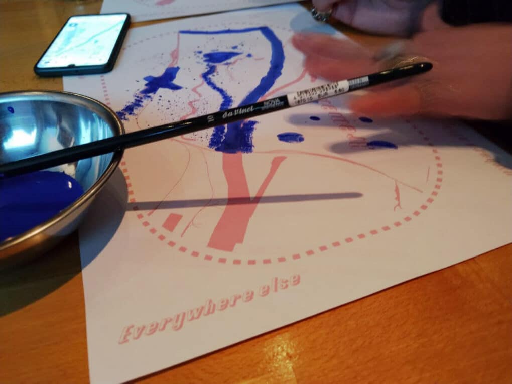 Color photograph of two hands over a desk; on the desk thereis a sheet with an pink basemap and overlaid drawings in blue, a bowl with more blue paint, and a smartphone with a digital map onscreen.