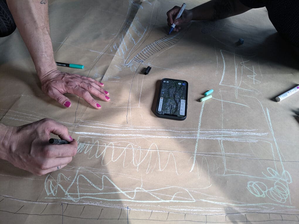 Hands of two people, one with pink nailpolish drawing with colored chalk on large sheet of brown paper. In the middle of the paper is an iPhone with a map onscreen.
