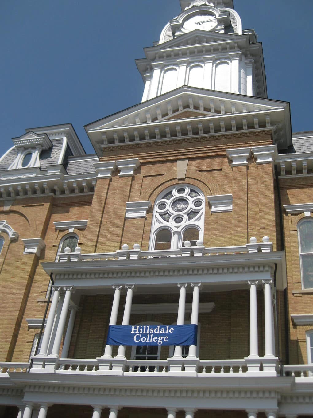 Brick college building with cupola, portico, and arched windows, with banner reading "Hillsdale College."