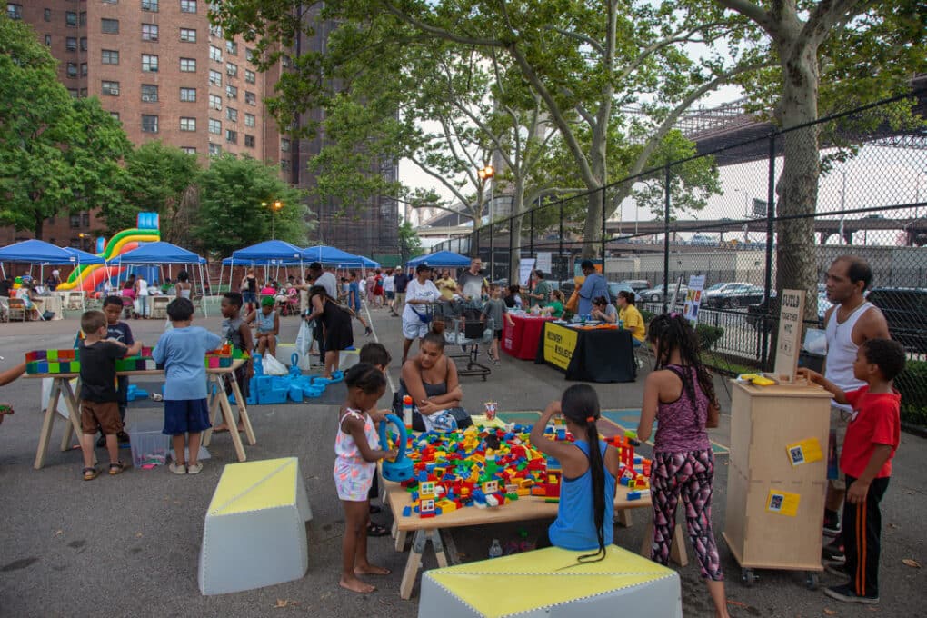 Color photograph of large group of mostly brown children at a Lego-building fair in tree-lined parkway beside housing-project towers.