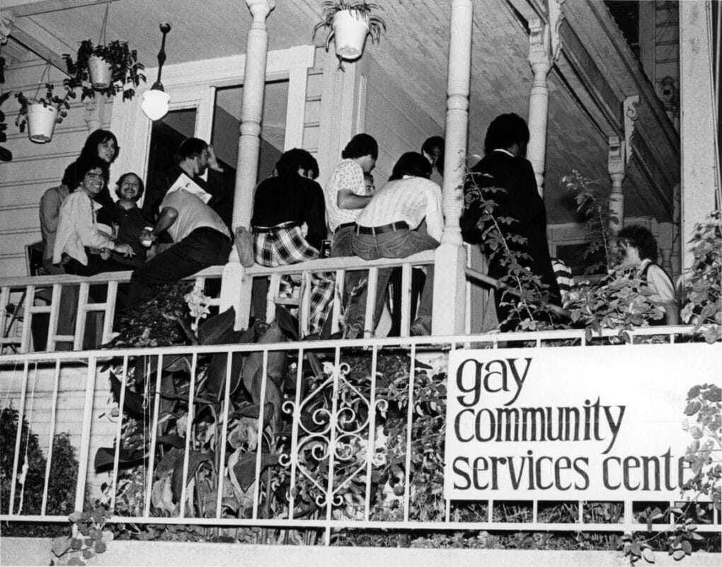 Black and white photograph of large group of casually dressed, long-haired, smiling white men sitting on railing of a large wooden house. A sign on the railing reads: "Gay Community Services Center."