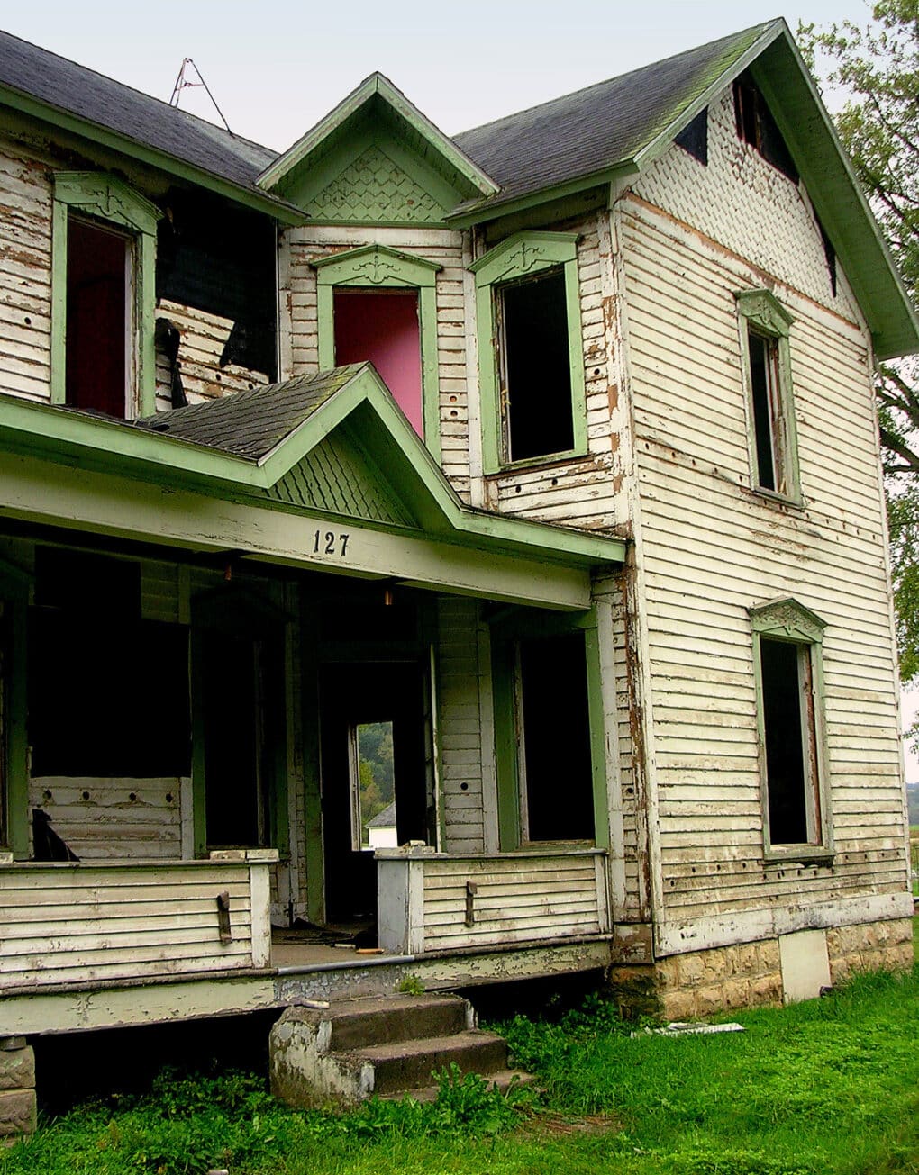 Color photograph of windowless, derelict two story wooden house with large porch.