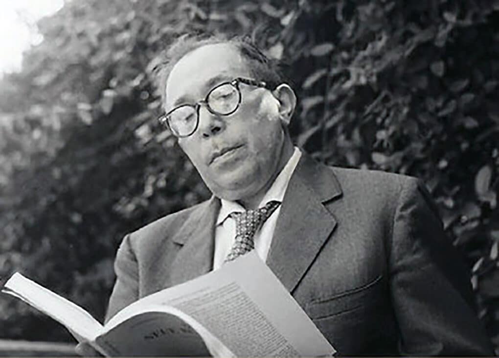 Black-and-white photo of white man in suit and tie reading a large book.