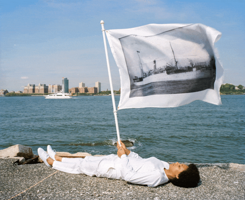 LaToya Ruby Frazier, <em>Holding flag laying at the edge of Pier 54 and the Hudson River.</em> On flag: US Army transport Buford, the “Soviet ark,” used to deport political radicals and other “undesirable” noncitizens from the U.S. to Russia in December 1919, broadside view, 1907, from <em>Pier 54: A Human Right to Passage,</em> 2014. 