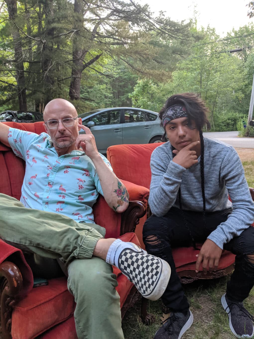 Two men, one middle-aged and bald, apparently white, and one young, with black hair in braids, apparently Native American, sitting side by side on red upholstered chairs in a yard with woods in the background. They look very much alike.