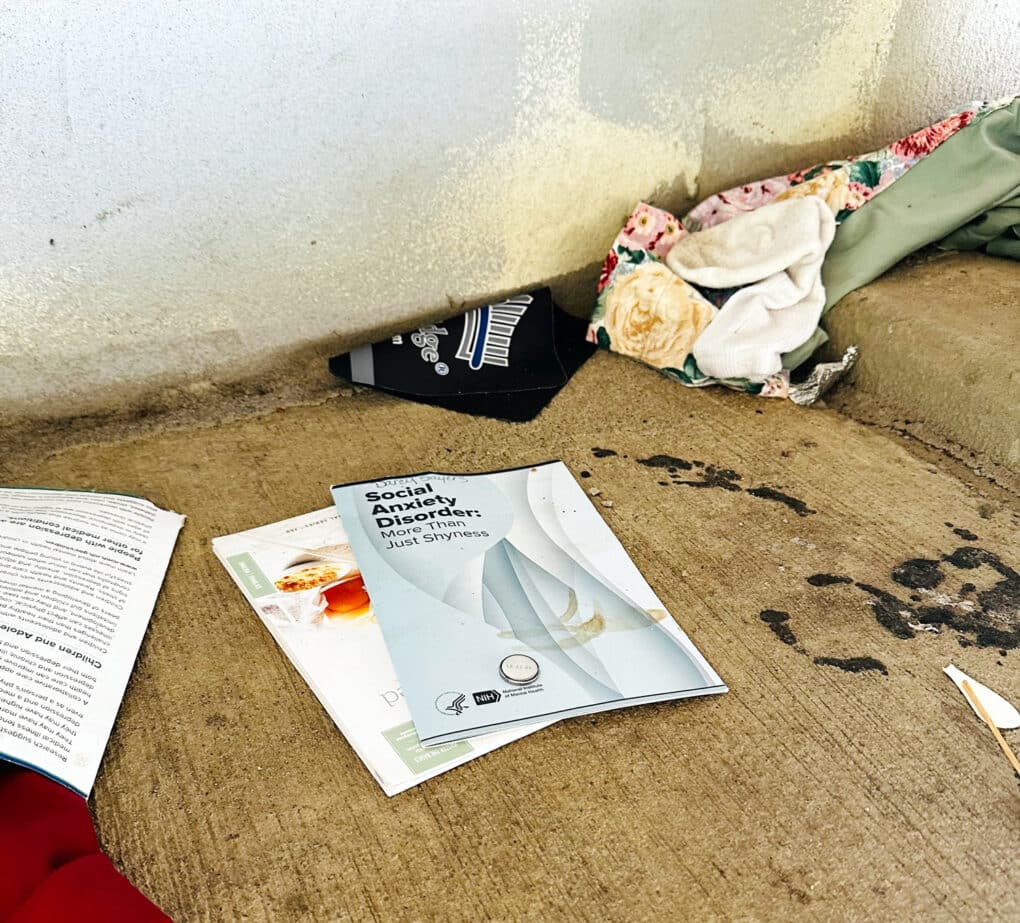 Color photograph of assorted clothes and papers left in a heap on a concrete walkway. Brochure at the front of image reads: "Social Anxiety Disorder: More Than Just Shyness."