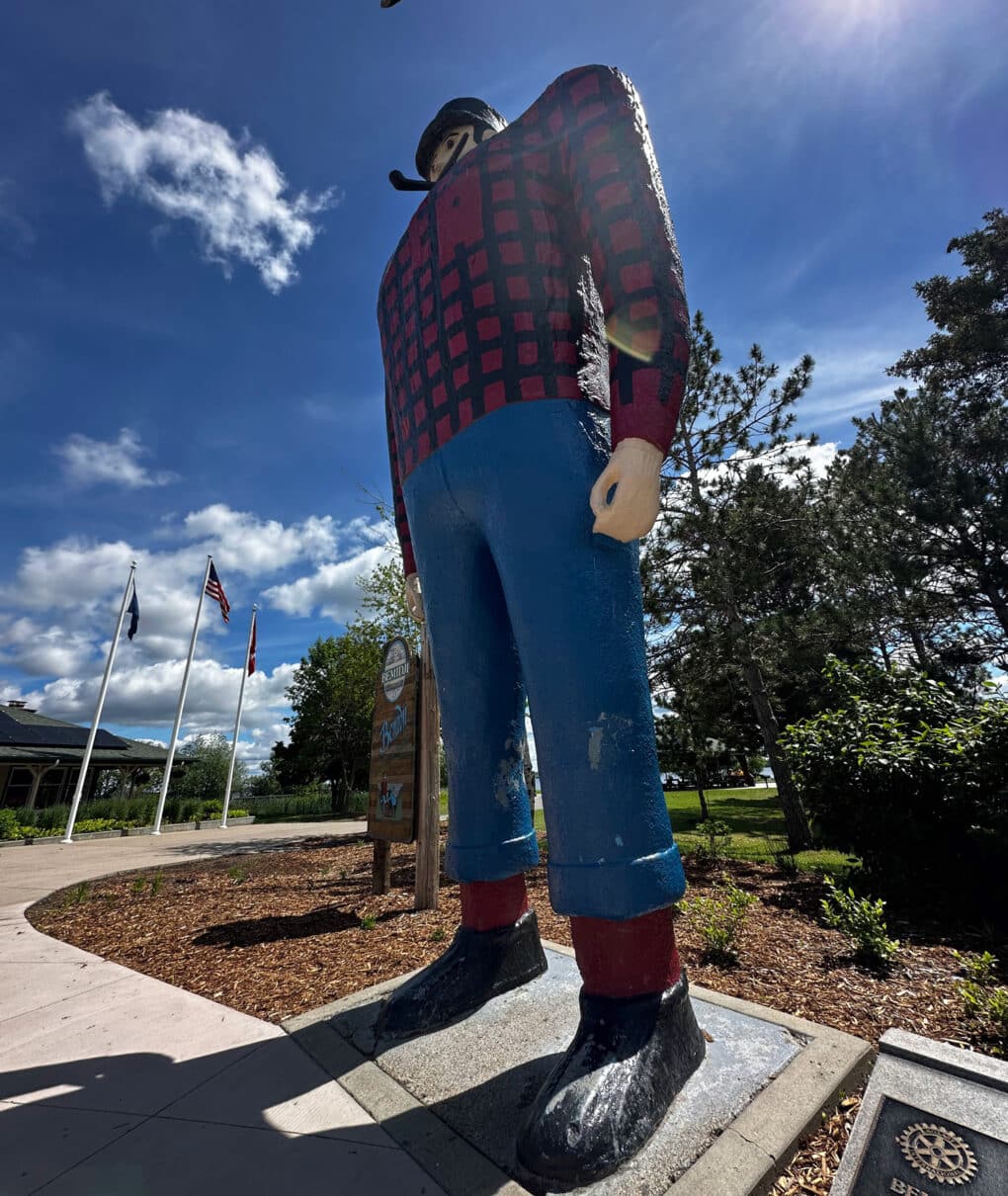 Color photograph of huge statue of man wearing blue pants, black, boots, and red-and-black lumberjack shirt, standing on a plinth with trees, blue sky, and American flag in background.