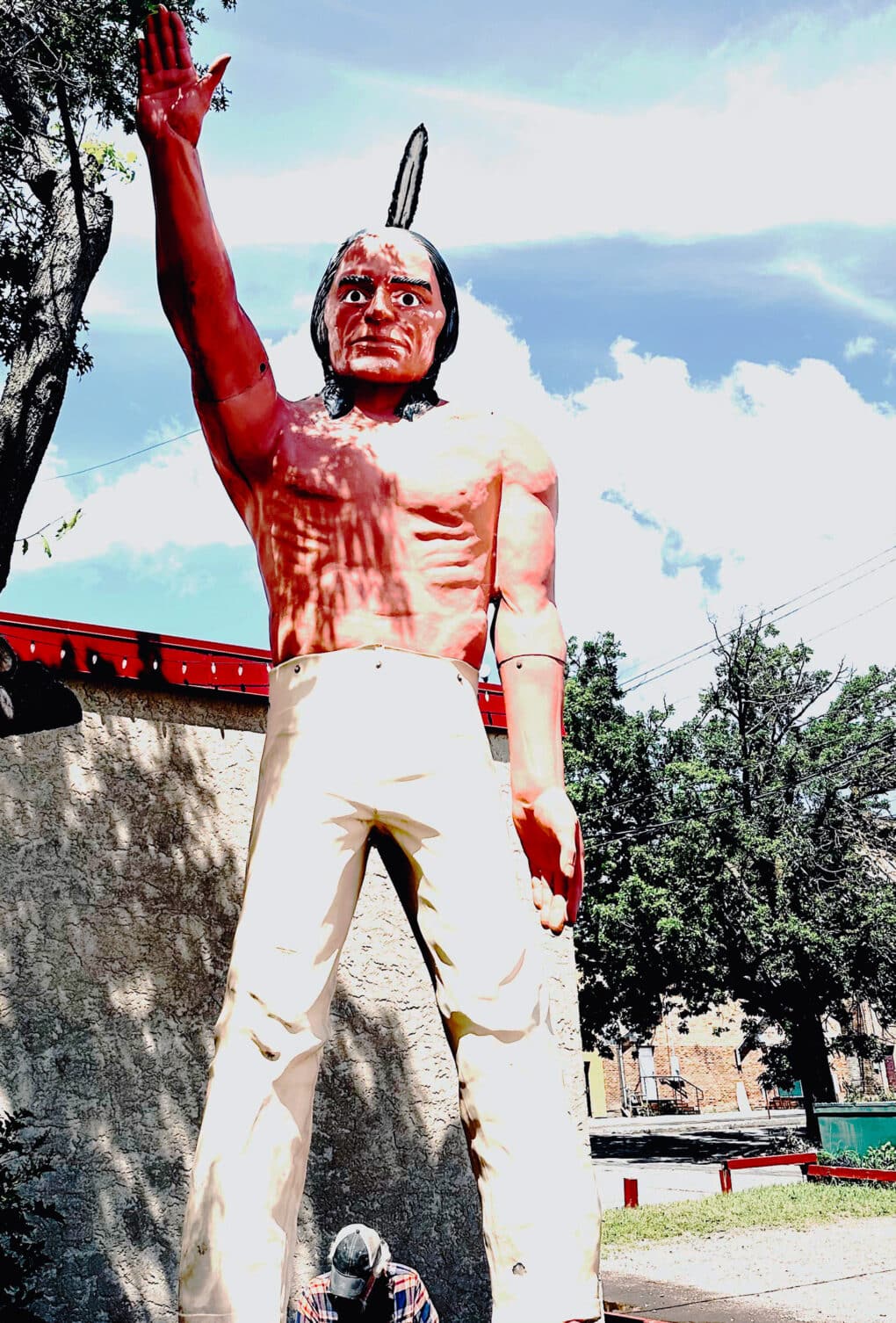 Monumental statue of red-skinned Indian man, shirtless, with an upright feather in his hair and a raised right arm.