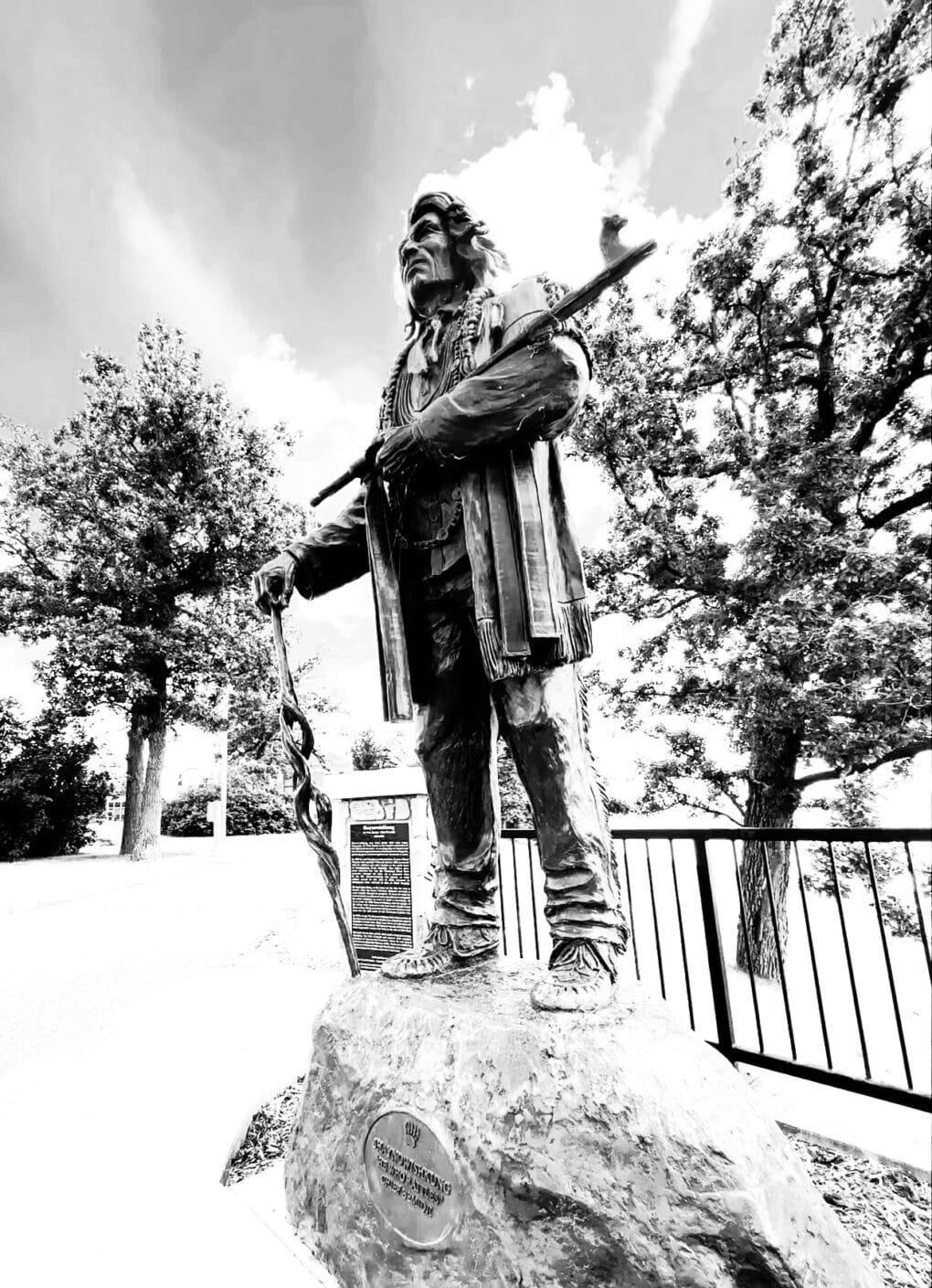 Black-and-white photograph of life-sized bronze of Native American man, wearing buckskins and moccasins and carrying a large pipe, standing on a rock-shaped plinth in a public park.