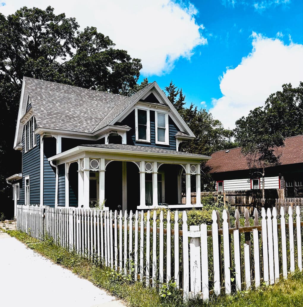 Color photograph of small wooden house on a corner lot, with simple gothic-style trim and a beat-up white picket fence.