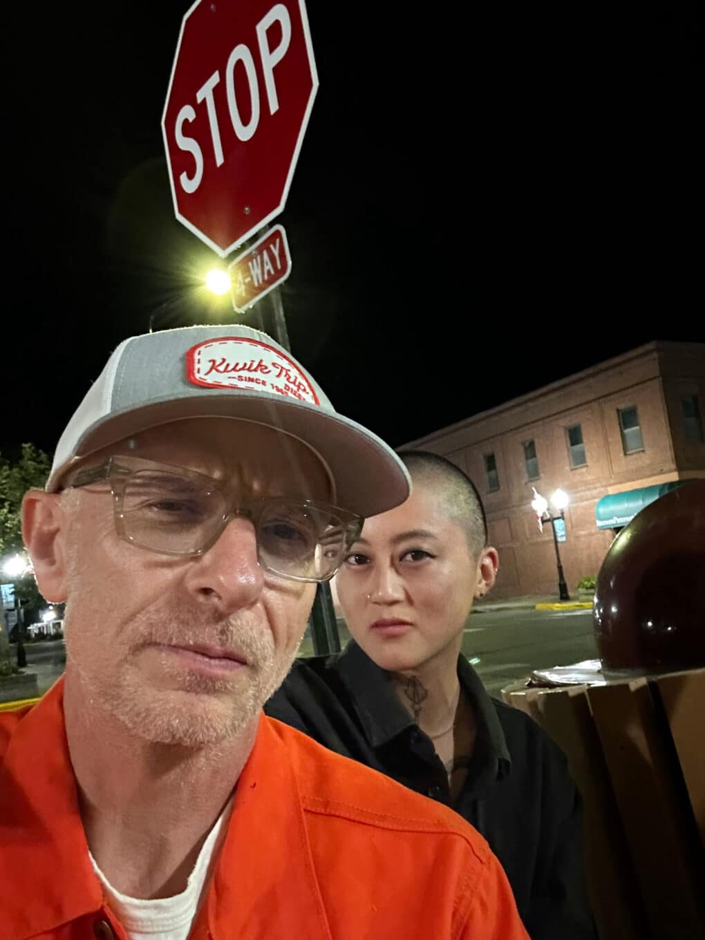 Color photograph of apparently white man in orange shirt and trucker hat, and Asian woman with shaved head, standing under a STOP sign. It is night, and a street lamp flares above their heads.