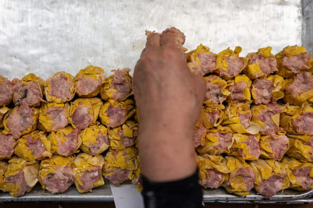 Color photo of stacks of dumpling wrappers stuffed with meat, uncooked, with a hand adding more to the pile.