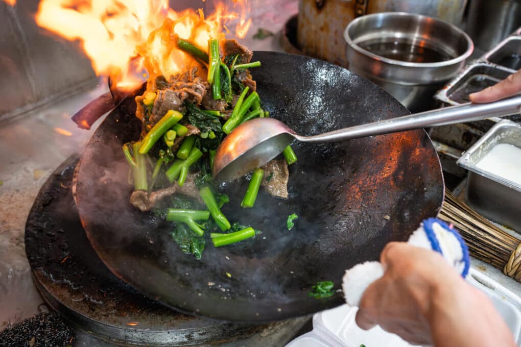 Greens and beef being stir-fried at high heat in wok, with flames. The cook is stirring the dish with a ladle.