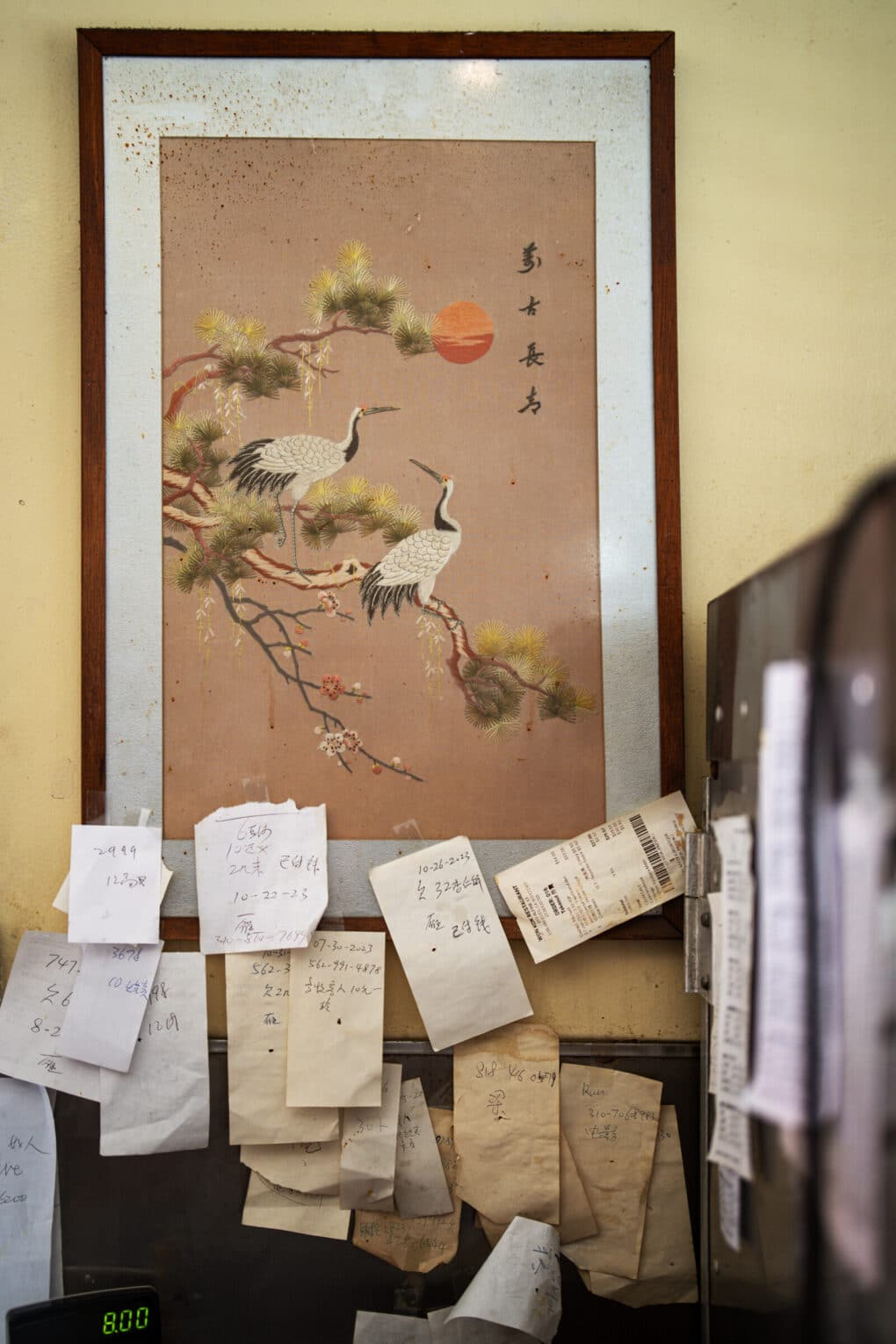 Apparently a photo of the counter area at Won Kok, with cash register visible, and many handwritten notes on scraps of paper, some yellowed with age, taped to a framed print of two birds. 