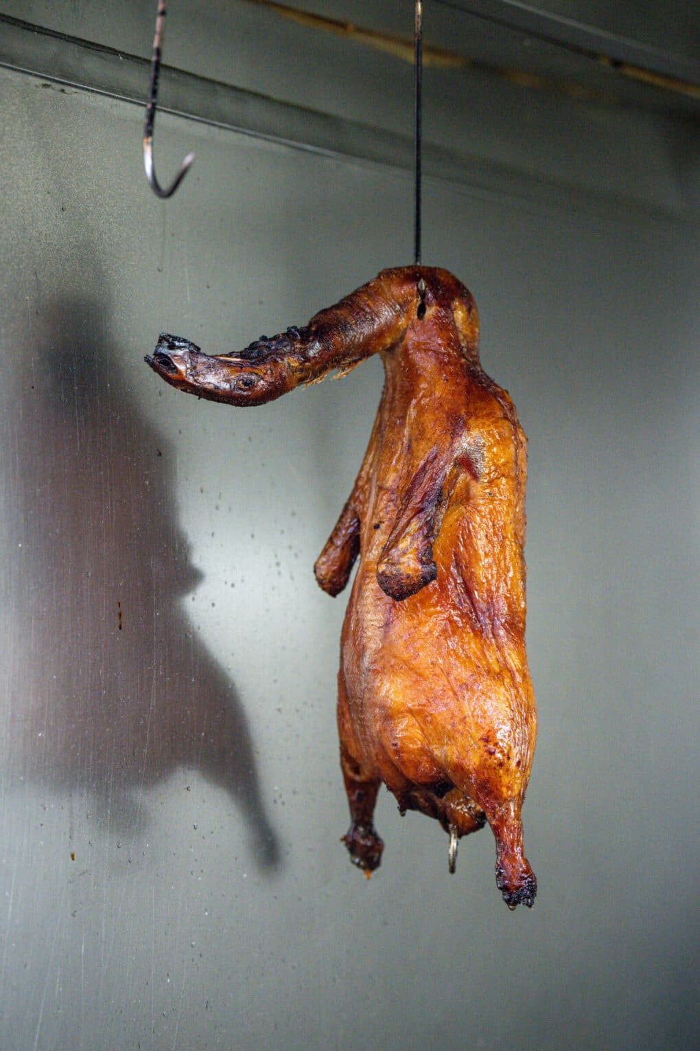 Preparing Peking duck in the Won Kok kitchen; a browned, marinated duck, head intact, is hanging from a metal hook. 