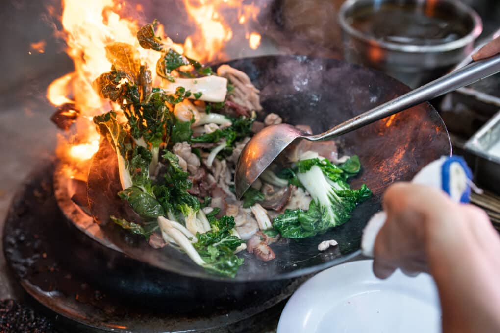 Beef and greens being cooked in wok over high heat, with flames. 