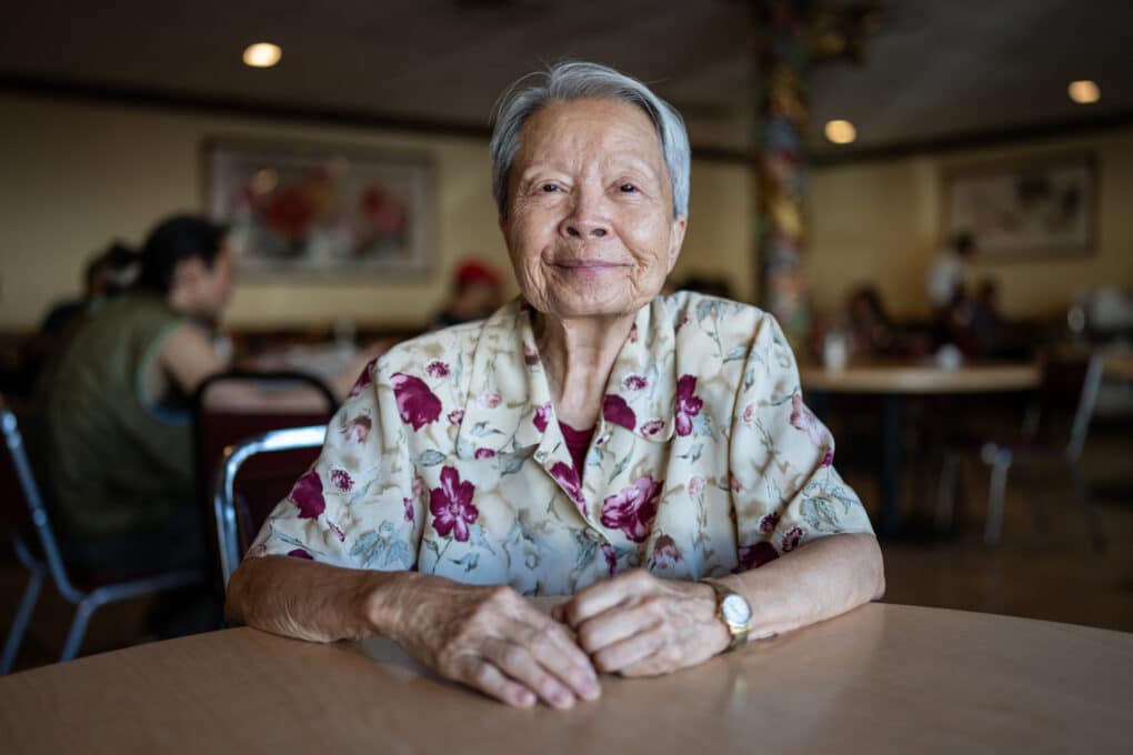 Sylvia Hom in the Wok Kok dining room, an older Chinese woman with short gray hair, smiling, wearing a blouse with flowers and a gold watch. 