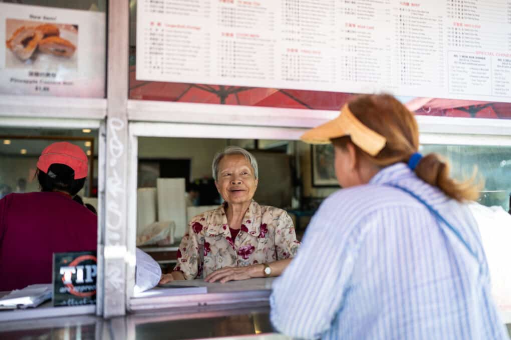 A Chinese woman with short gray hair is smiling at a customer ordering food from a takeout window.