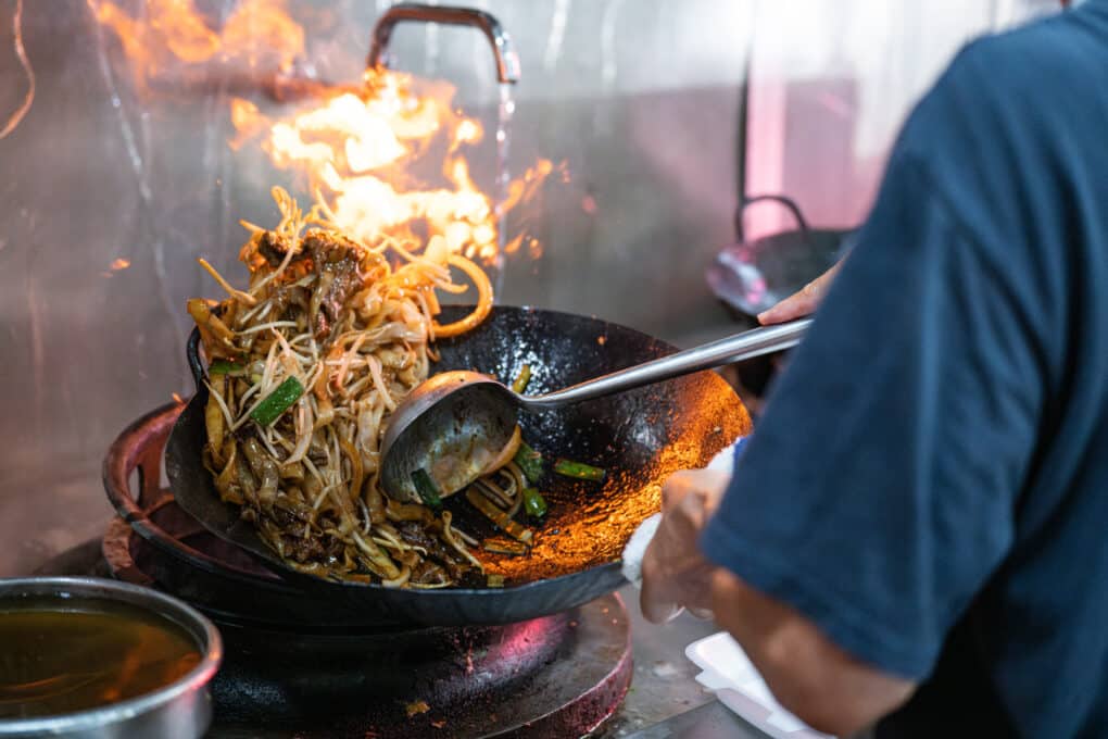 A cook is using a ladle to stir beansprouts and meat in a very hot wok with flames. 