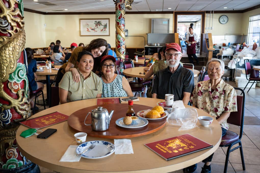 A group of five people of mixed age and ethnicity are sitting around a round table, smiling, with food remnants and tea pot and Won Kok menus.