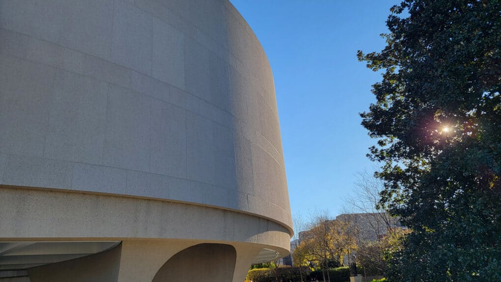 Color photograph of curved concrete wall of brutalist building, against bright blue sky.