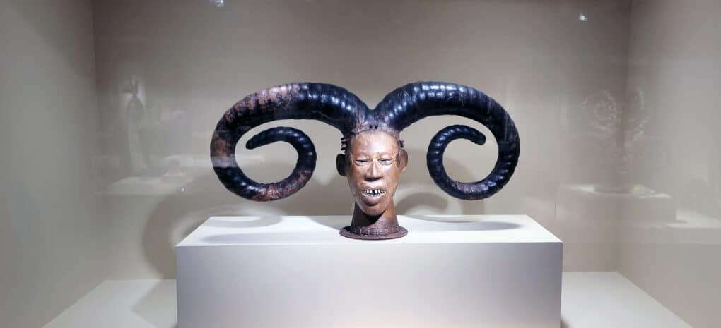 Color photograph of African bronze sculpture of double-horned human head, in a museum vitrine.