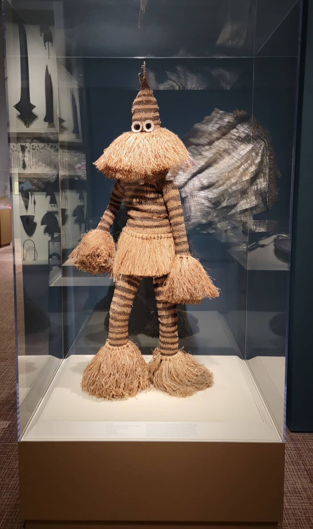Color photograph of masked African figure made of raffia in museum vitrine.