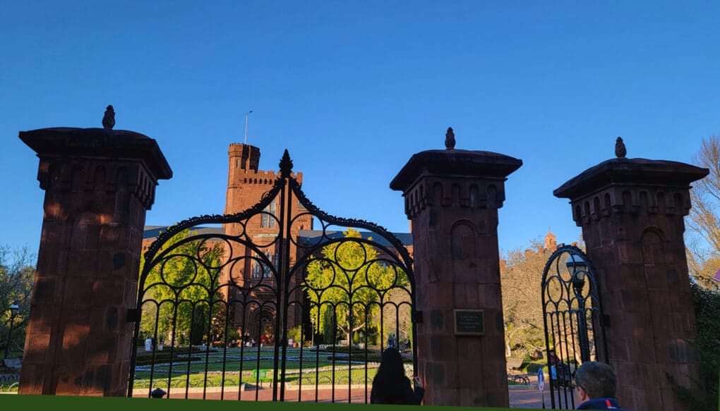 Color photograph of red-brick turretted building, with brick pillars and wrought-iron gate in the foreground, against bright blue sky.