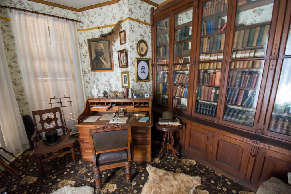 Color photograph of wallpapered room with rolltop desk, glass-fronted bookcase, framed photograph of Frederick Douglass, and other framed portrait photographs.