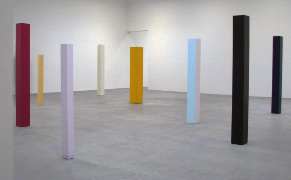 Color photograph of eight tall colored plinth-like sculptures in white-cube gallery space.
