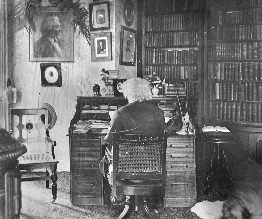 Black-and-white photograph of elderly man with seen from the back, seated at a desk with large bookcase to his right.