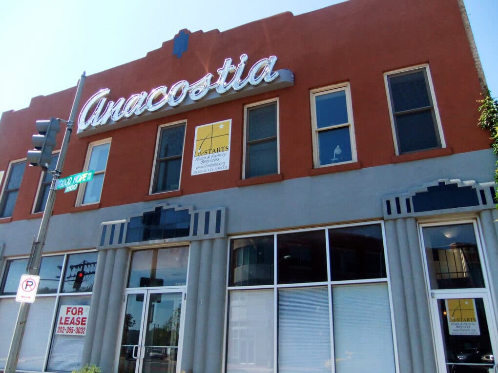 Color photograph of two-story brick building with neon sign reading ANACOSTIA and "For Lease" sign in storefront window.