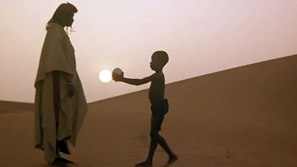 Color film still of woman and small boy in silhouette, against a sand dune with the sun low on the horizon. The boy is handing the woman an ostrich egg.