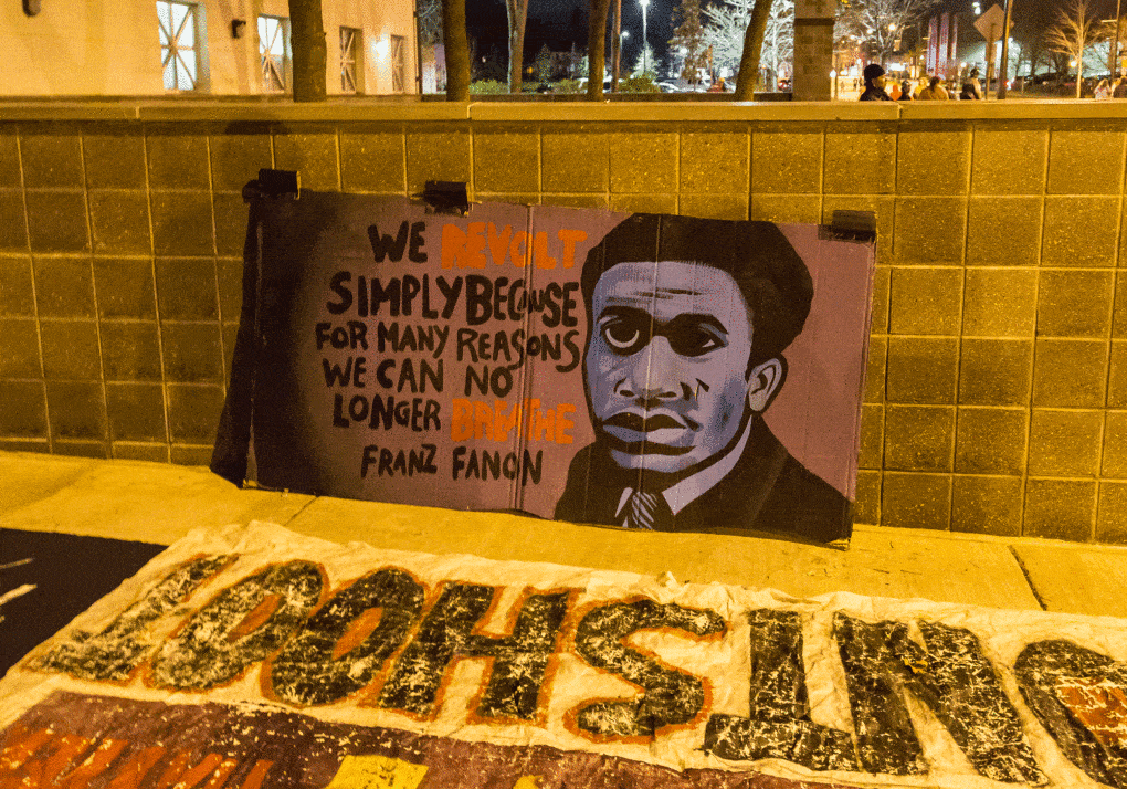 Protest at the Minneapolis Police Department Fourth Precinct following the police shooting of Jamar Clark, November 2015. The banner quotes Frantz Fanon.