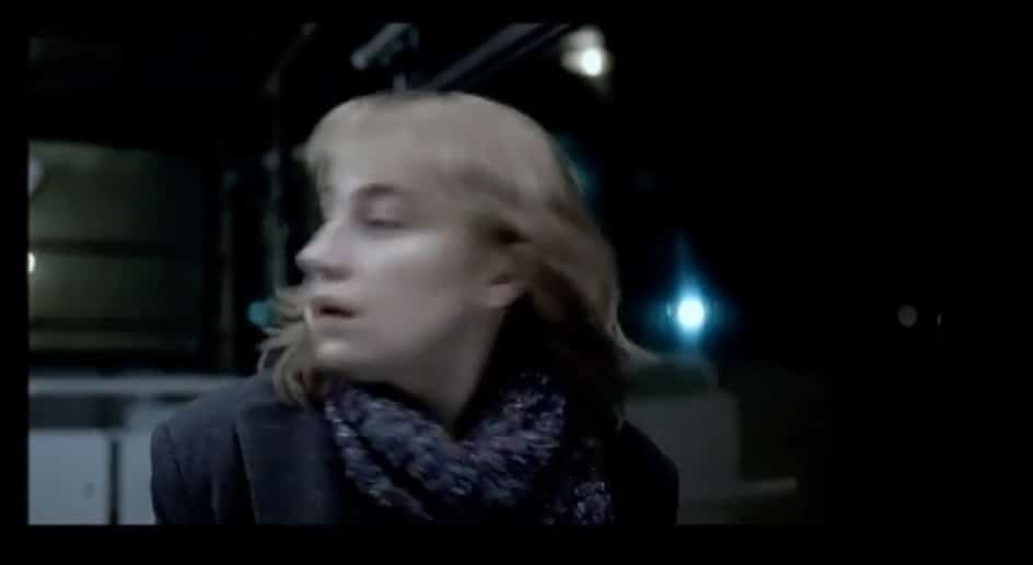 Color screenshot showing young blonde woman in winter clothes turning her head against a dark background on a subway train.