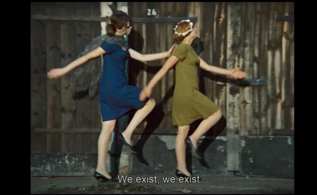 Color film still of two young white women in 1960s-style dresses skipping in unison, seen in profile, against a wooden fence. The subtitle reads "We exist, we exist..."