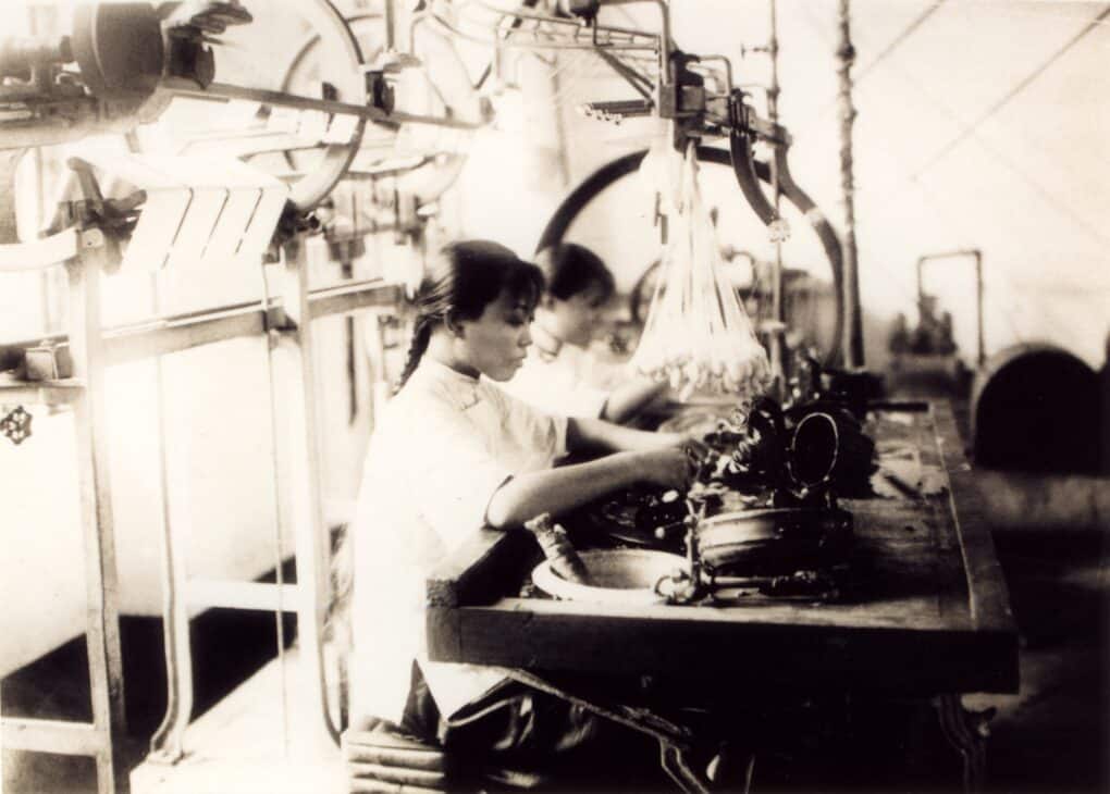 About three women are working intently at a metal bench; a cluster of small cocoons hang near their faces, and there is machinery directly behind.