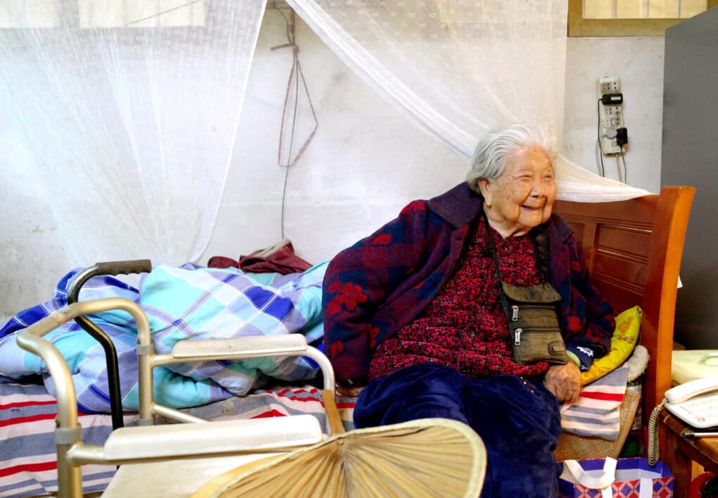 An elderly Chinese woman is sitting on the edge of her bed, smiling broadly.