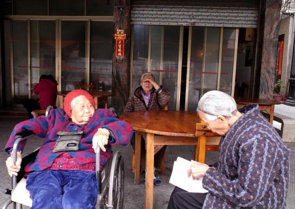 Three older women are sitting near a table, in conversation. One is in a wheelchair, another is browsing a book.
